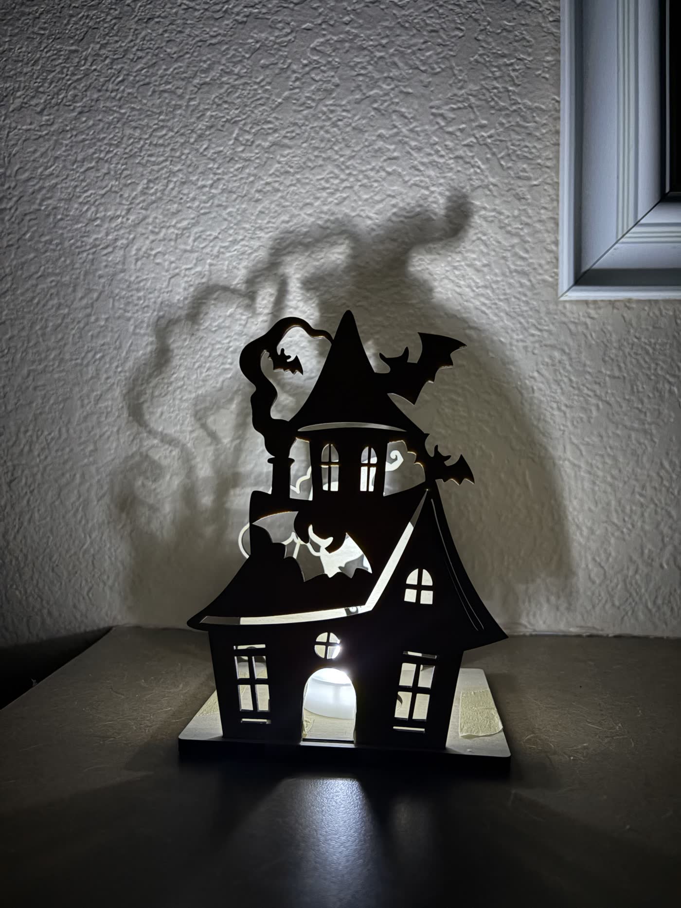 Wooden Halloween lantern shaped like a witch’s house with bats, casting a house-and-smoke shadow on the wall.